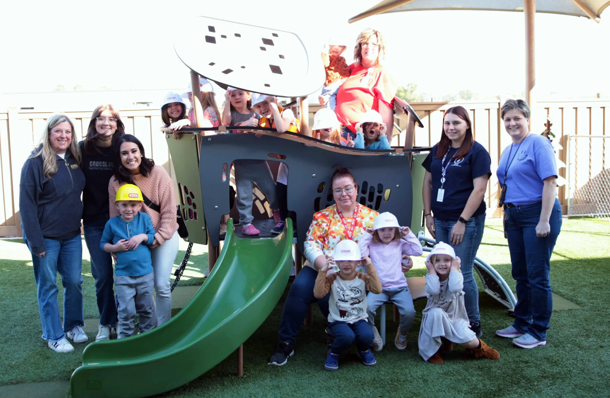 A group of adults and children pose and smile together around a playground structure with a slide on a sunny day. Some children wear hats and costumes, and everyone looks happy.
