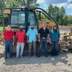 Five people stand in front of a large bulldozer on a construction site, with trees and a blue sky in the background. Three men wear hats, and everyone is dressed in casual attire.