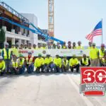 A large group of construction workers in safety gear pose in front of a building site, holding a beam. An American flag waves in the background. A “30 Years Building Arkansas” badge is in the lower right corner.
