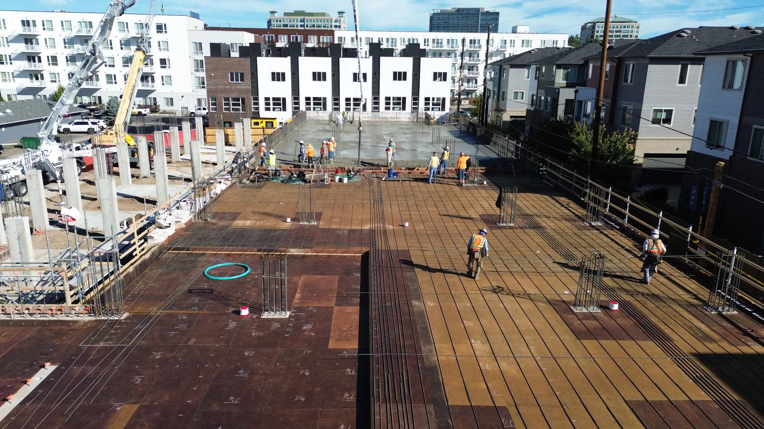 A construction site with workers preparing and pouring concrete on a building’s foundation, surrounded by apartment buildings and equipment under a clear blue sky.