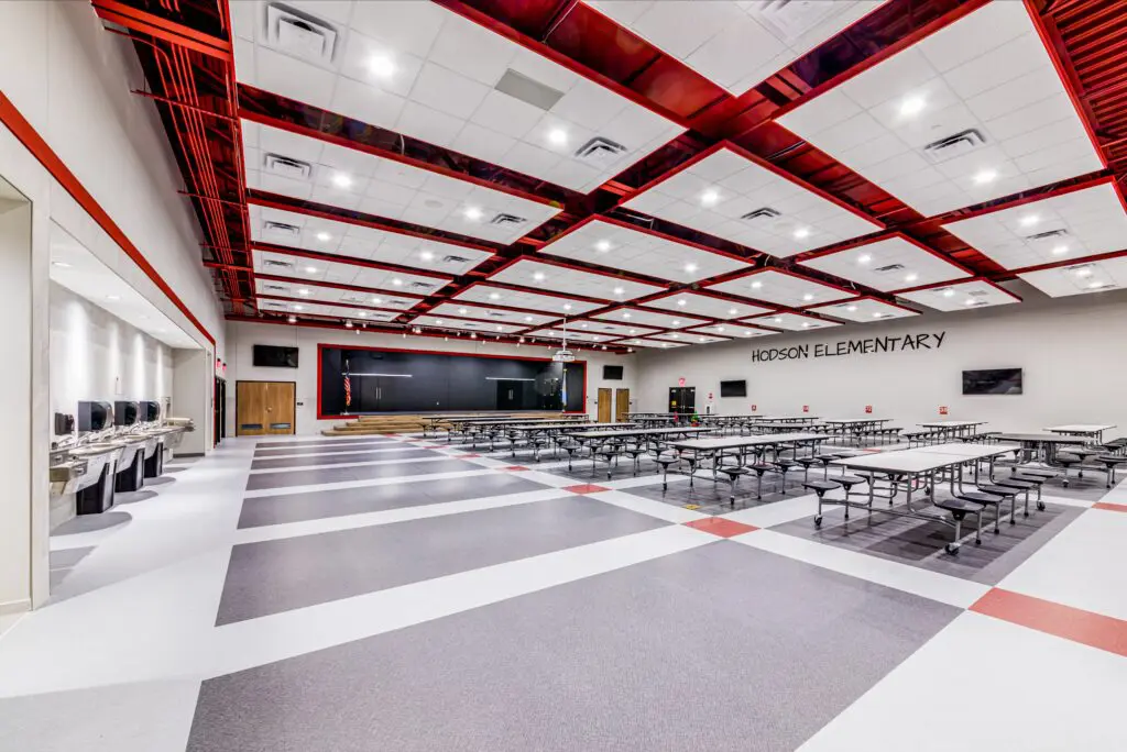 A spacious, modern cafeteria with long tables and benches, a stage at one end, and “Hodson Elementary” written on the wall. The ceiling features red beams and bright lighting.