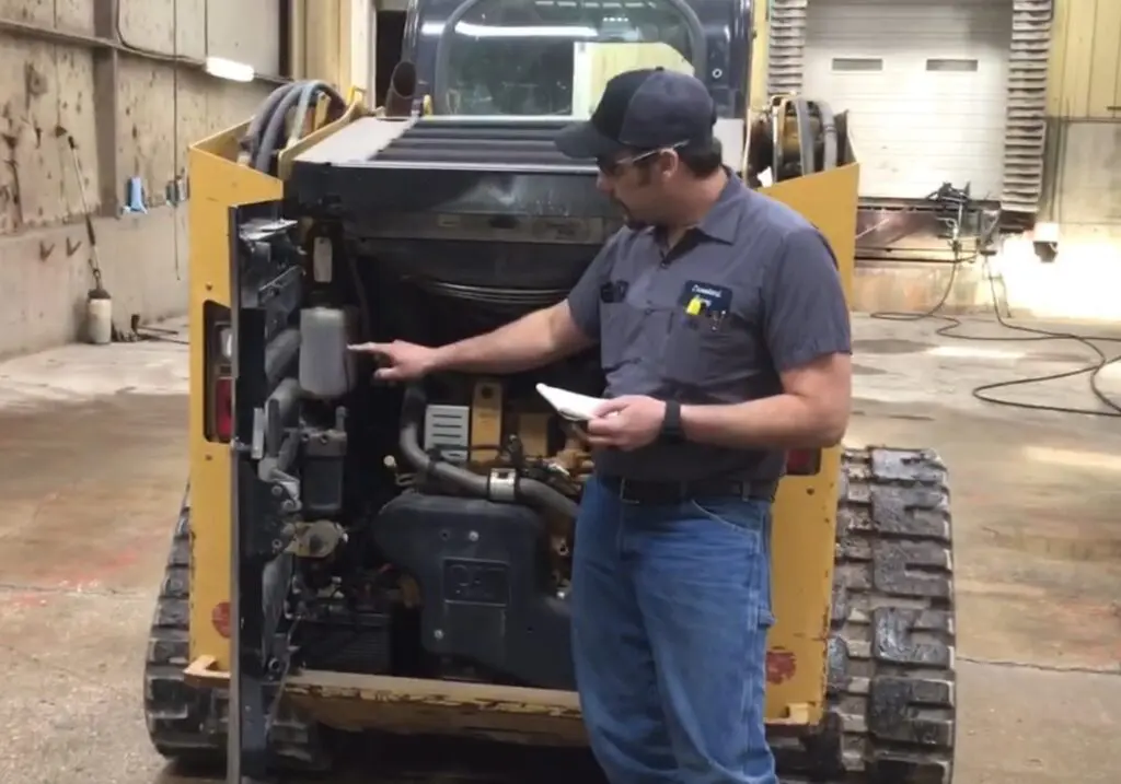 A man in work clothes points at machinery components inside the open rear compartment of a yellow tracked skid-steer loader in a workshop. He is holding a notepad and inspecting the equipment.