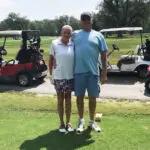 A smiling couple stands arm-in-arm on a golf course, with golf carts and greenery in the background. The woman wears a white shirt and floral shorts; the man wears a blue shirt and shorts with a cap and sunglasses.