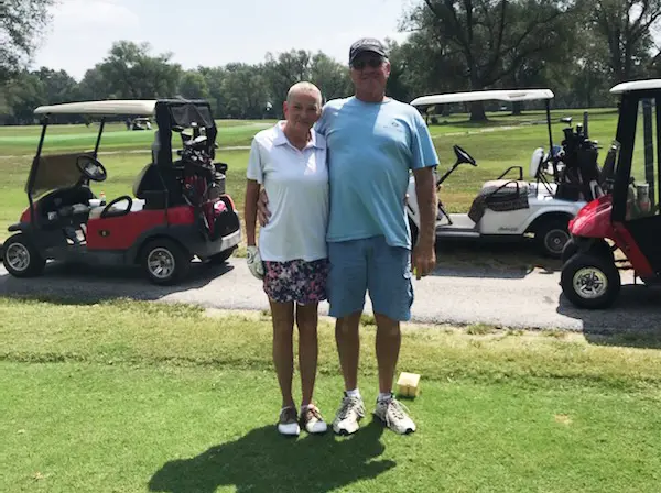 A smiling couple stands arm-in-arm on a golf course, with golf carts and greenery in the background. The woman wears a white shirt and floral shorts; the man wears a blue shirt and shorts with a cap and sunglasses.