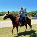 An older man in a cowboy hat waves while riding a brown horse with a small child sitting in front of him. They are outdoors on a grassy area with a road, cars, and trees in the background under a clear blue sky.
