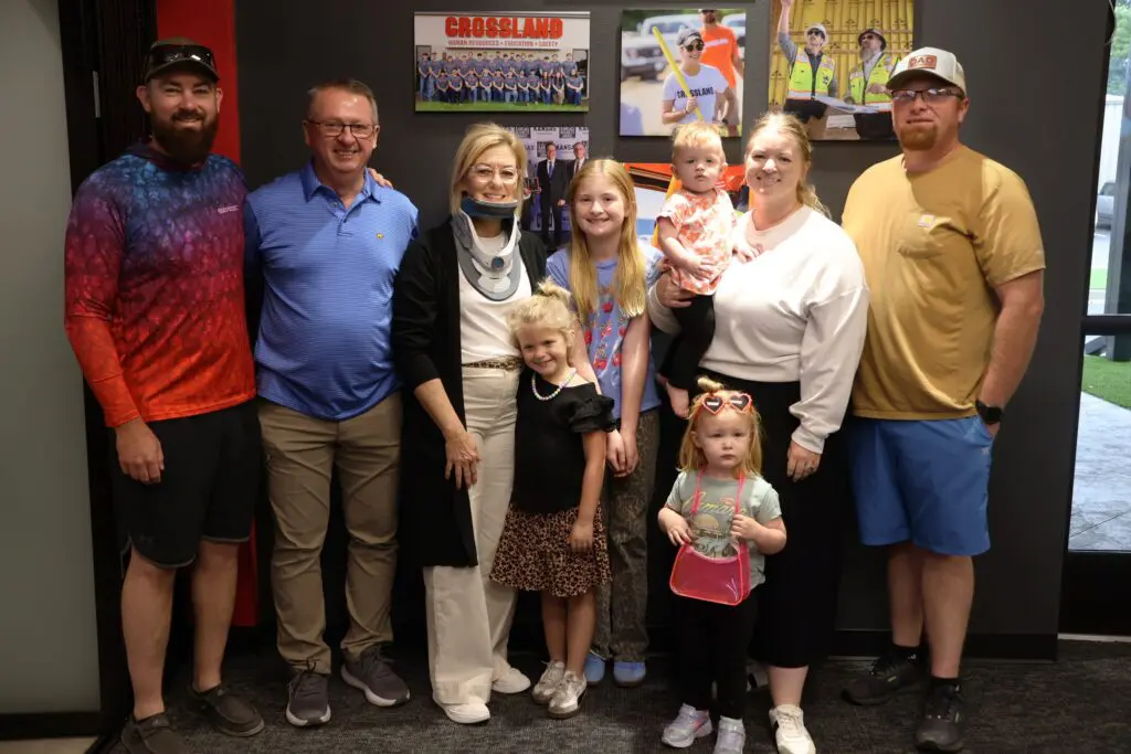 A group of eight adults and children stand together indoors, smiling at the camera. One woman in the center wears a neck brace. Photos and posters are displayed on the wall behind them.