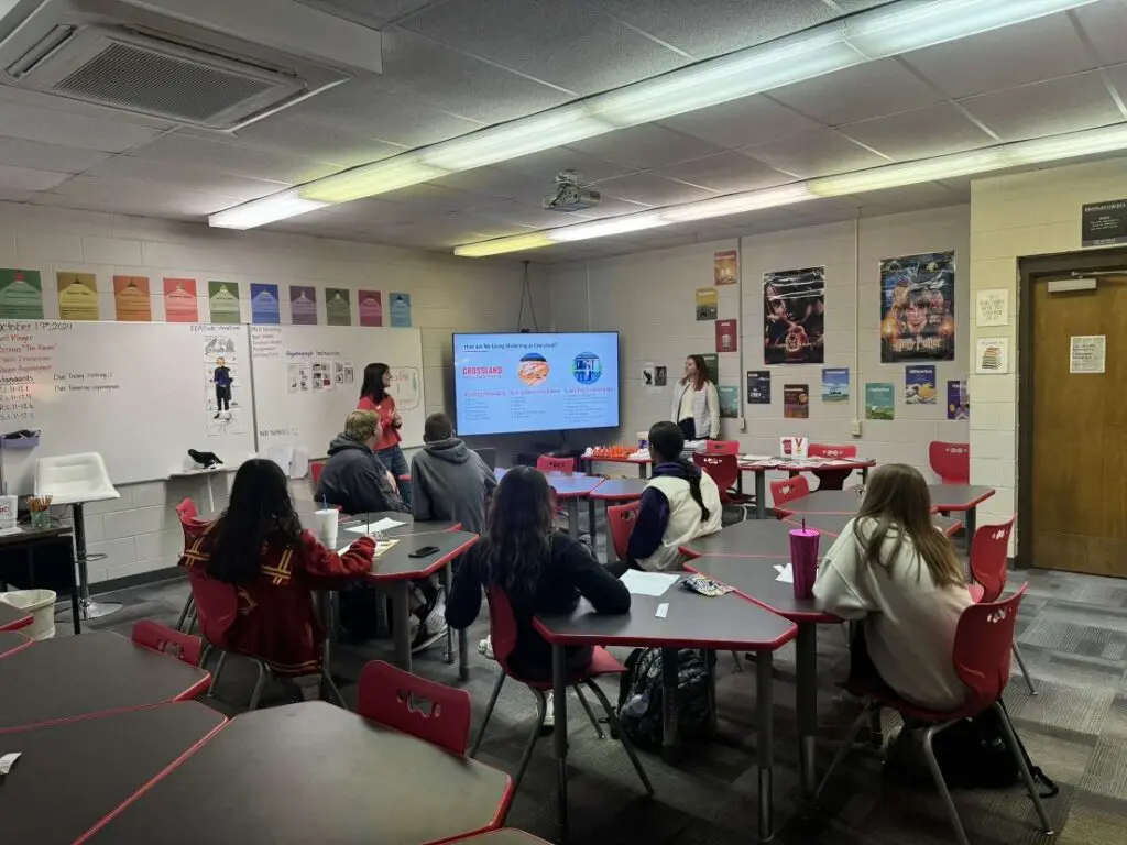 A classroom with students seated at red tables, facing a large screen displaying a brain diagram. Two presenters stand at the front, while posters and colorful signs decorate the walls.