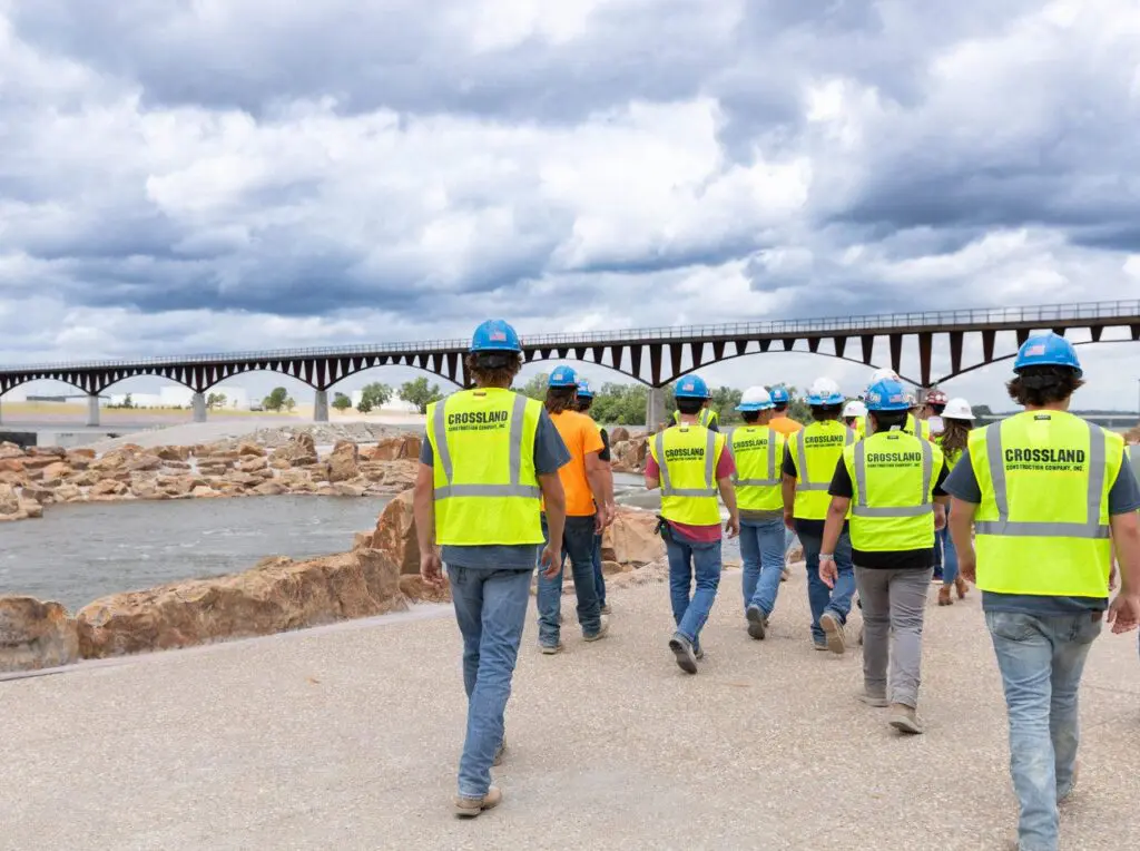 A group of construction workers in yellow vests and blue helmets walk near a river, with rocks and a long bridge in the background under a cloudy sky.