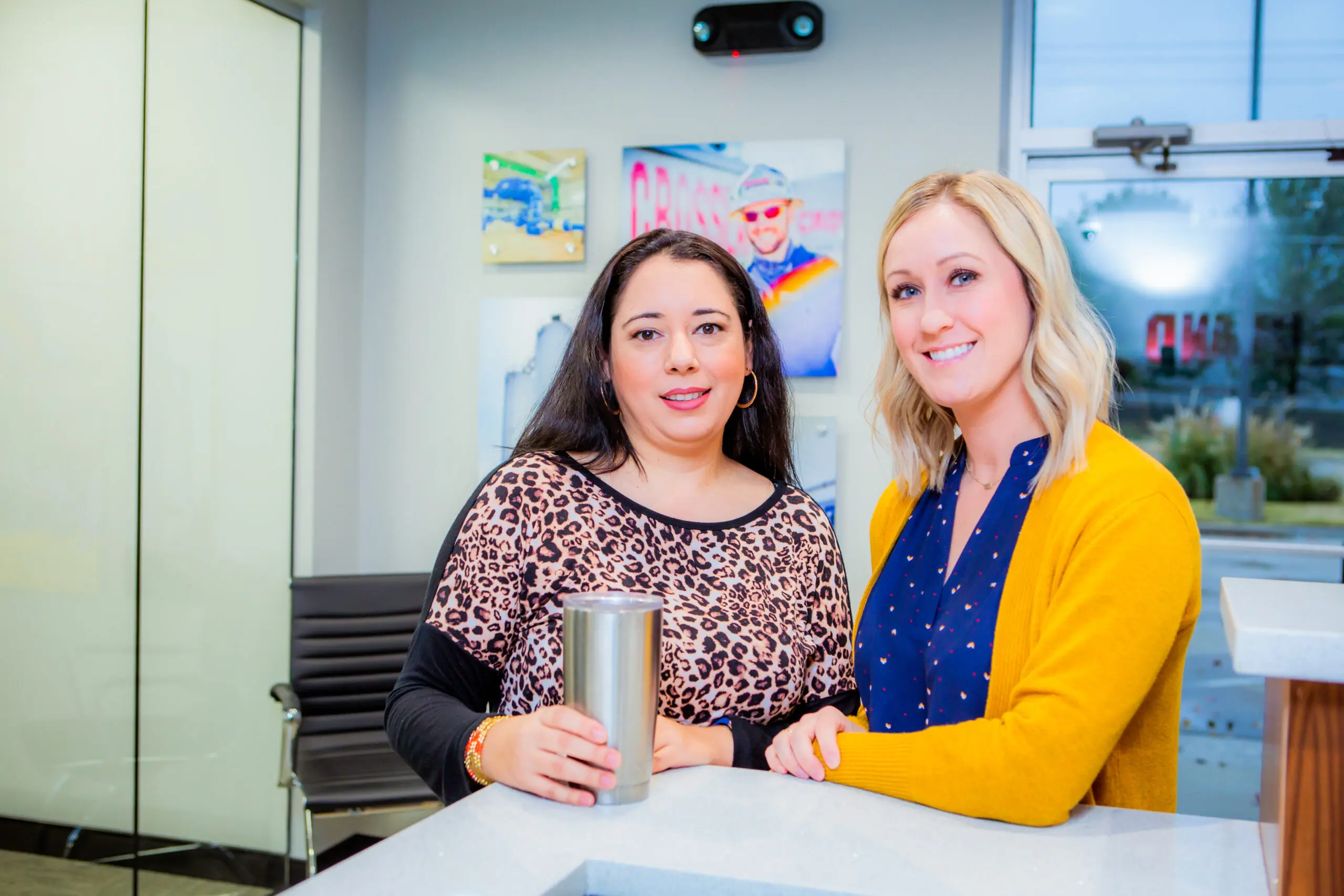 Two women stand side by side indoors, smiling at the camera. One has dark hair and wears a leopard-print top, holding a tumbler. The other has blonde hair and wears a yellow cardigan over a blue shirt.
