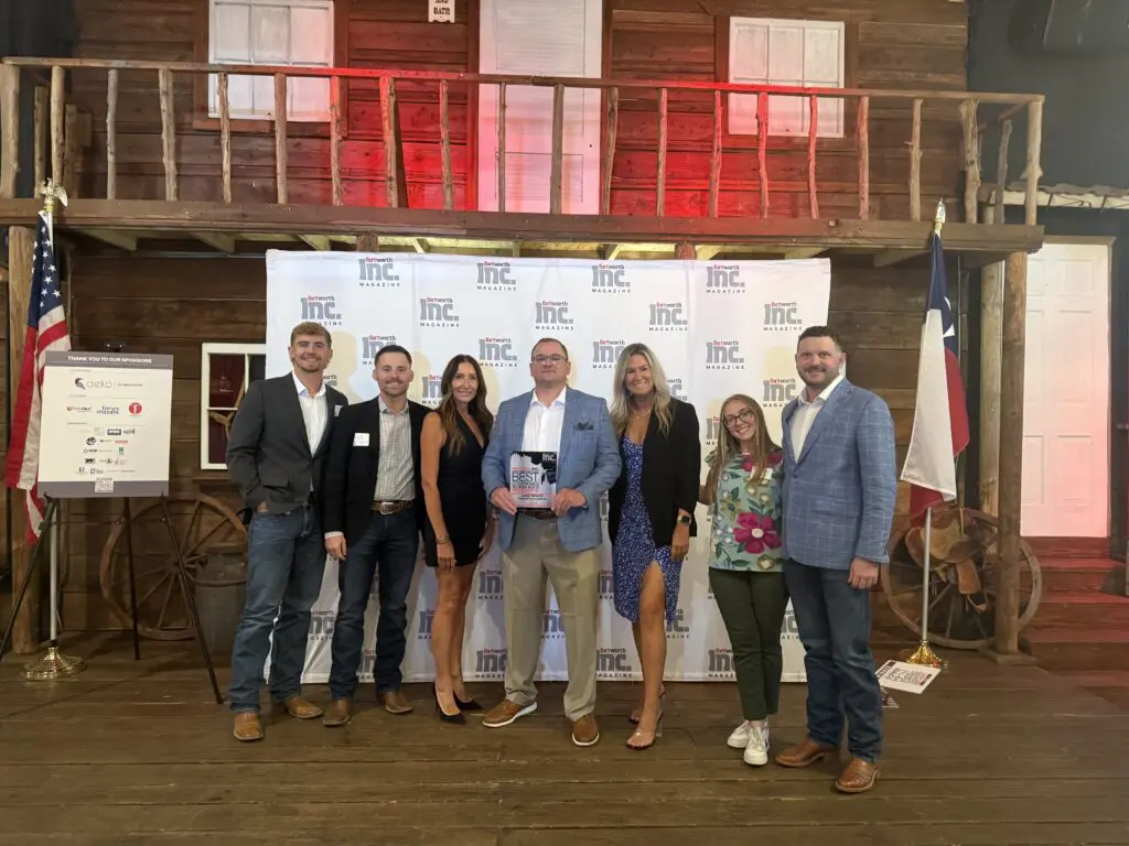 Seven people pose together on a wooden stage in front of an Inc. banner, with two U.S. flags on either side. One person in the center holds a clear award. The setting resembles a rustic barn.