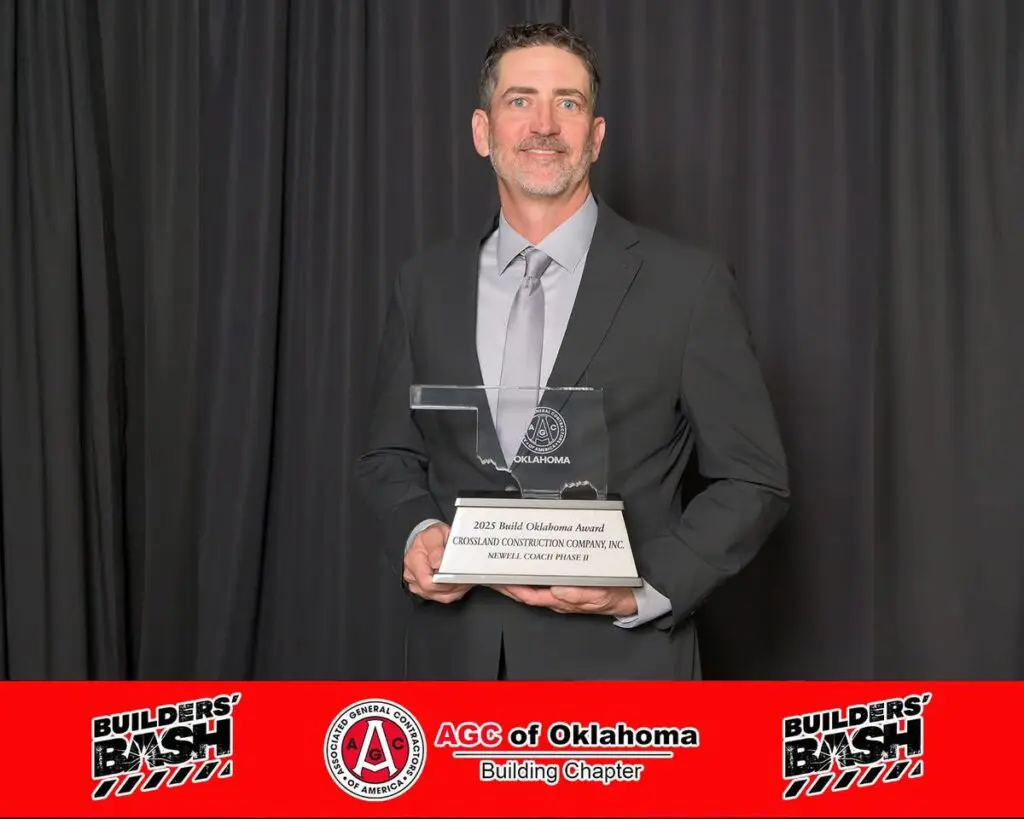 A man in a suit smiles while holding a clear plaque award. He stands in front of a dark backdrop with a red banner at the bottom displaying "AGC of Oklahoma" and "Builders' Bash" logos.