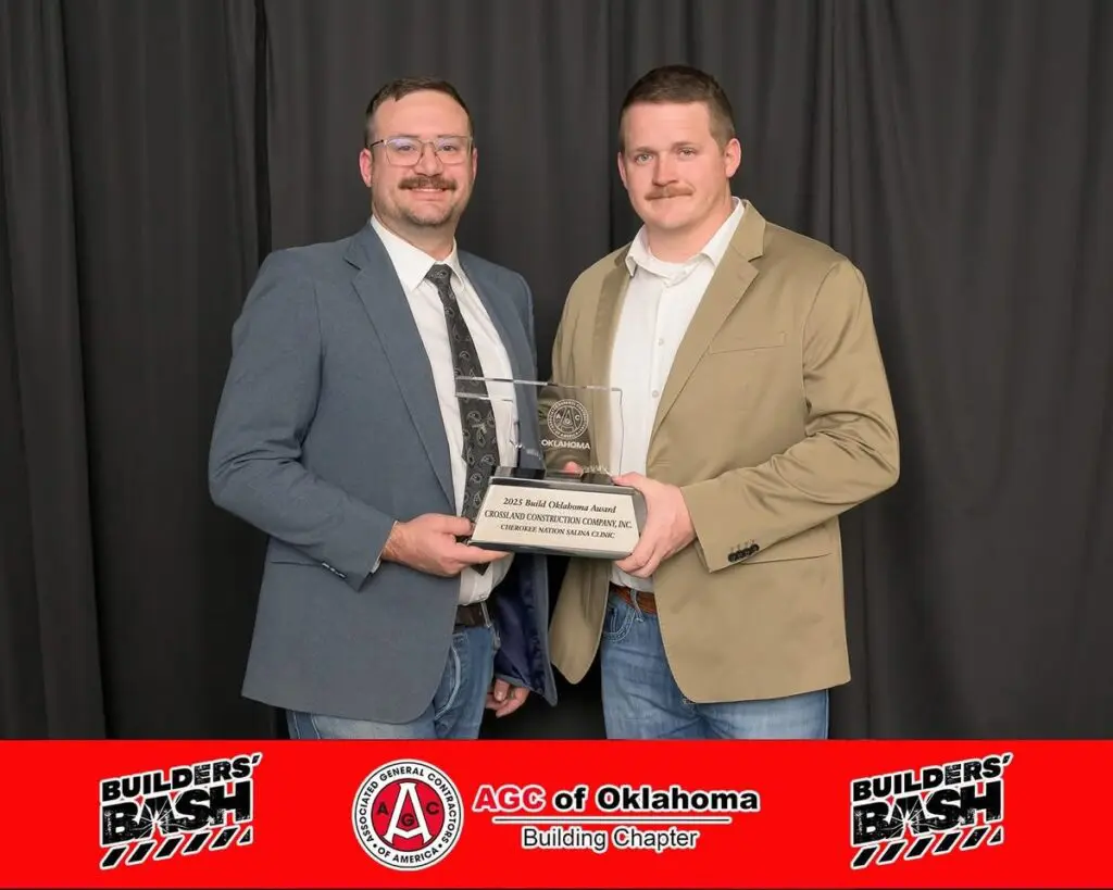Two men in suits stand together holding a glass award plaque. Behind them is a black curtain. At the bottom, a red banner reads "Builders’ Bash, AGC of Oklahoma, Building Chapter.