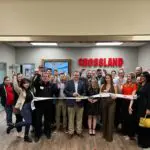A large group of people stand inside an office, smiling and holding a ribbon for a ribbon-cutting ceremony. A sign reading "CROSSLAND" is on the wall behind them. Several people wave at the camera.