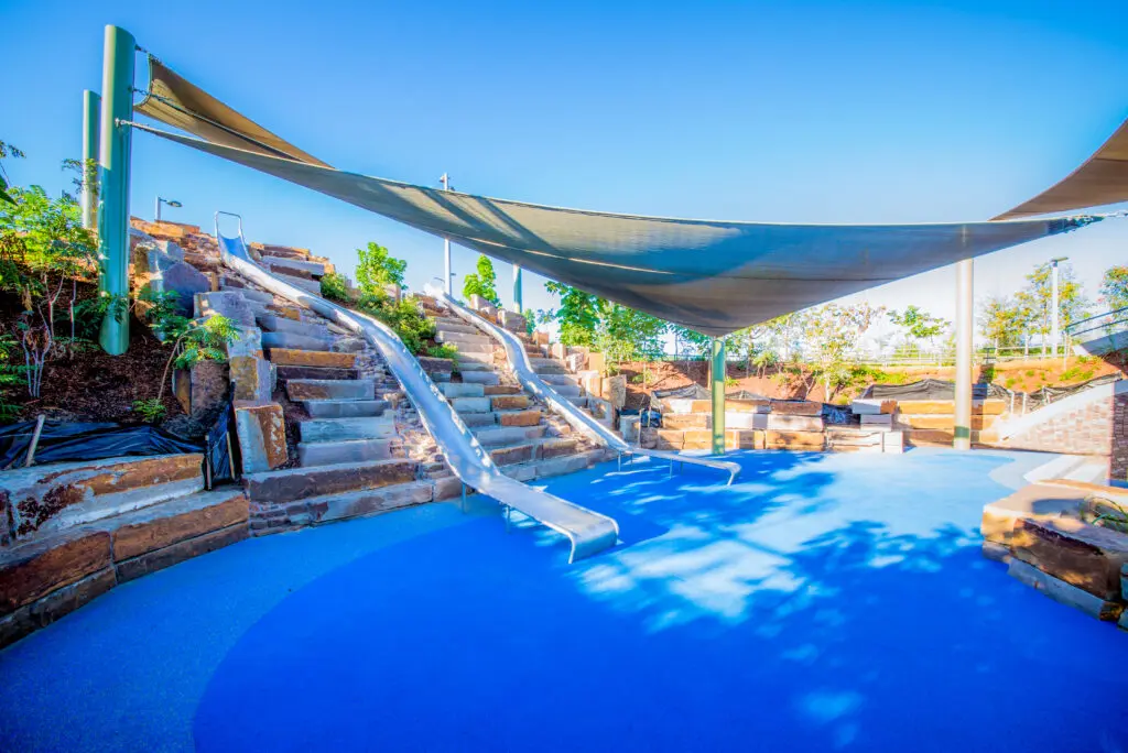 A vibrant playground features three metal slides built into a rocky hill, with blue rubber flooring and large shade sails overhead. Greenery and trees surround the area under a clear, sunny sky.