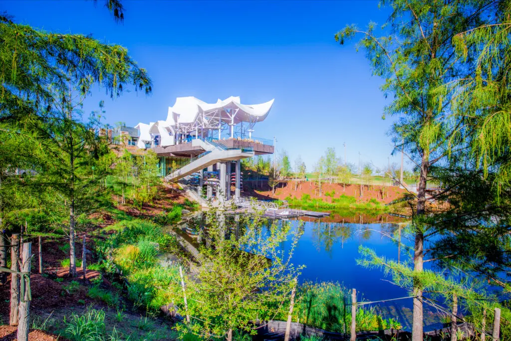 A modern, white-roofed pavilion stands on stilts above a green landscape, overlooking a calm pond surrounded by trees and vegetation, under a clear blue sky.
