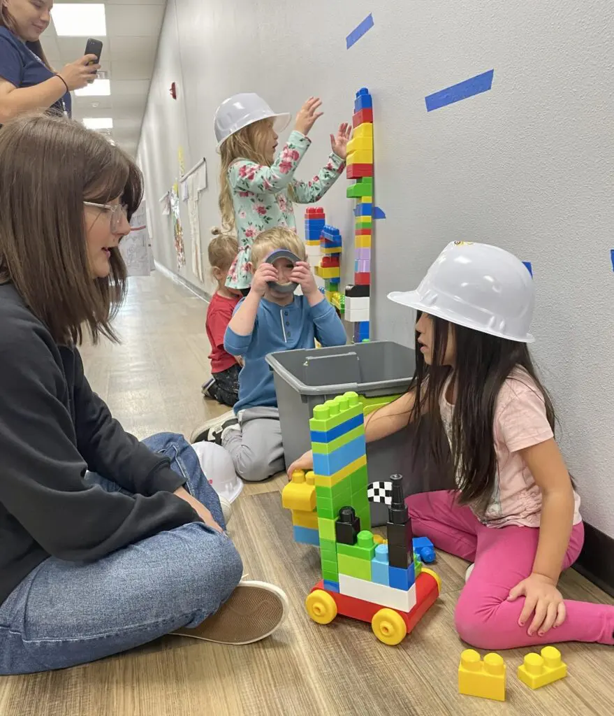 Several young children, some wearing toy hard hats, play with large colorful building blocks along a hallway. An adult sits nearby, watching and engaging with the children as they build.