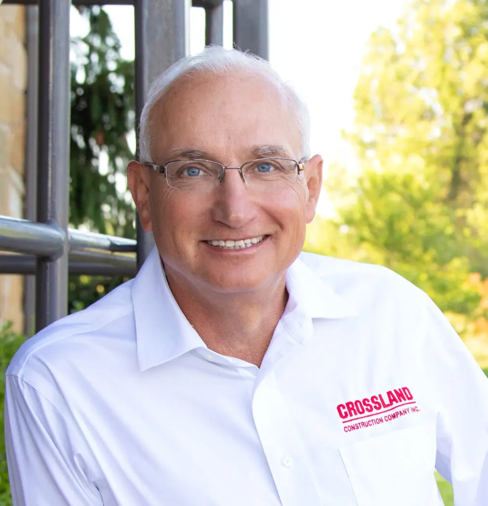 Smiling older man with short gray hair and glasses wearing a white shirt with "CROSSLAND Construction Company Inc." logo, standing outdoors near metal railings and greenery in the background.