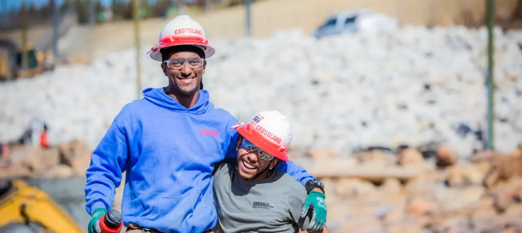 Two construction workers wearing hard hats and safety gear smile and pose together at a rocky outdoor job site, with one worker’s arm around the other.