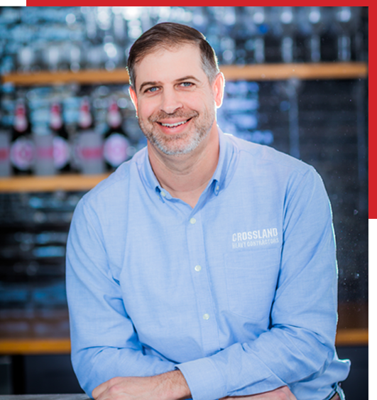 A smiling man with short brown hair and a beard, wearing a light blue button-up shirt with "CROSSLAND" embroidered on it, sits at a counter with bottles and glasses blurred in the background.