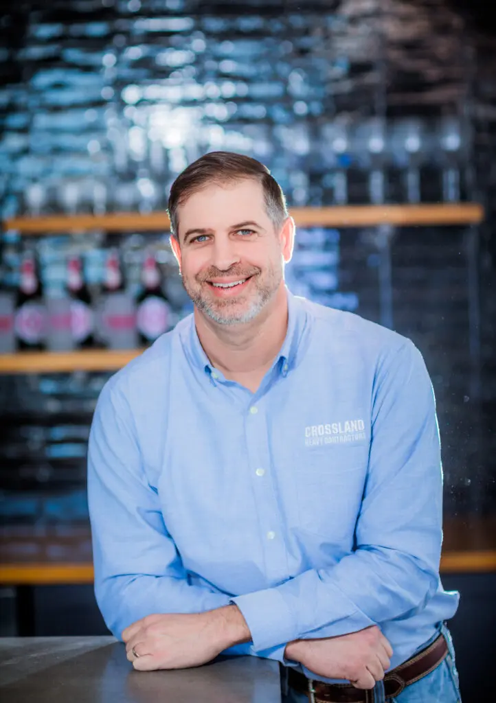 A man with short brown hair and a beard, wearing a light blue button-up shirt with "CROSSLAND HEAVY CONTRACTORS" on it, smiles while leaning on a counter in front of shelves with bottles and glasses.