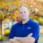 A man with a shaved head stands outdoors in front of autumn trees, wearing a blue collared shirt with "CROSSLA" embroidered on it, arms crossed, looking at the camera with a neutral expression.