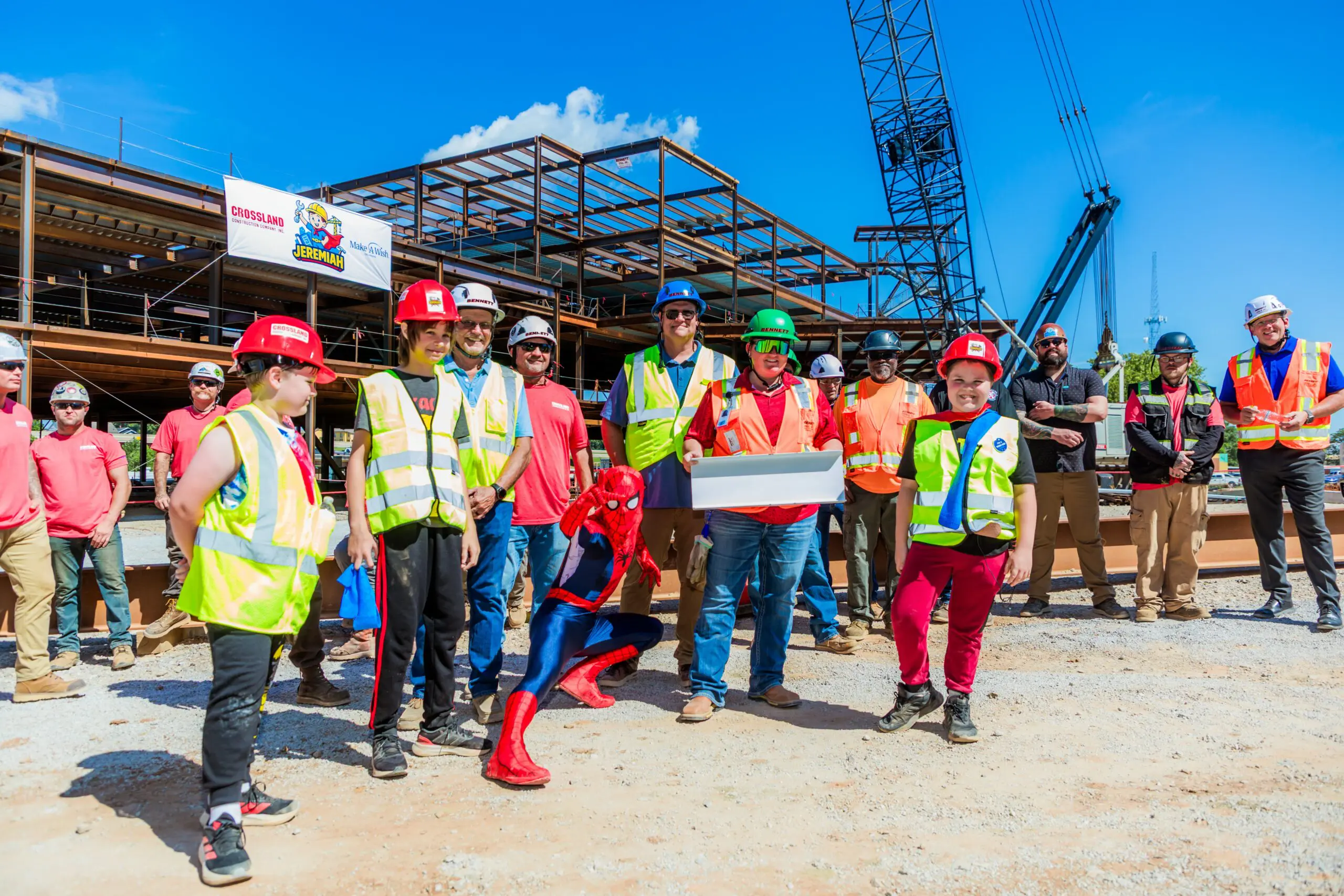 A group of construction workers in safety gear pose at a building site with steel framework; a person dressed as Spider-Man crouches in front, and one worker holds a white box. A crane and blue sky are in the background.