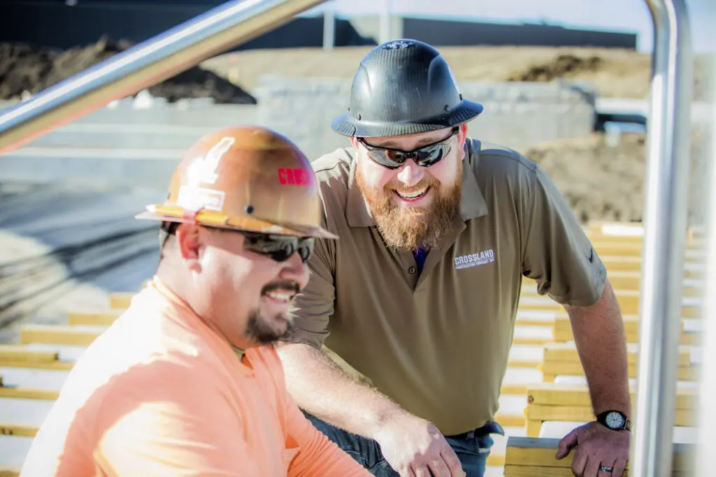 Two construction workers wearing hard hats and sunglasses smile and talk outdoors. One wears an orange shirt, the other a brown shirt and black hard hat. Wooden planks and a construction site are visible in the background.