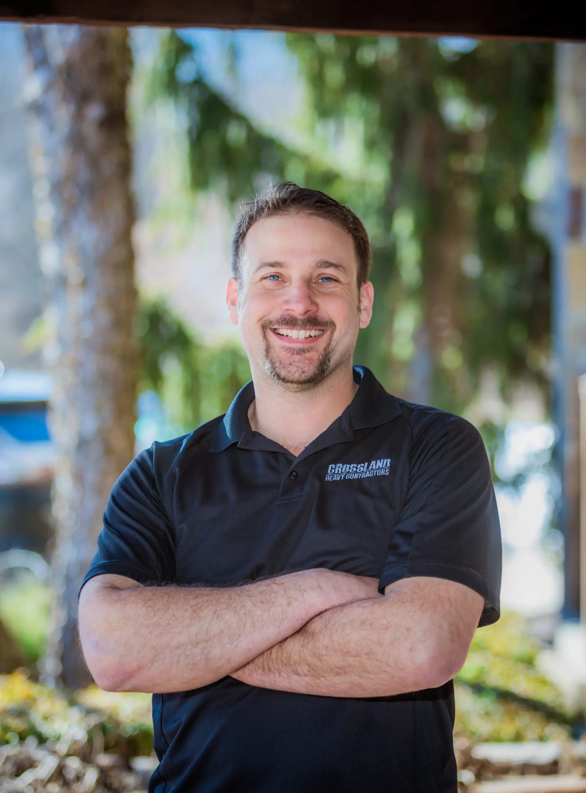 A man with short brown hair and a beard smiles with his arms crossed, standing outdoors in front of trees. He wears a black polo shirt with a small “CRISSLANE CONSTRUCTION” logo on the chest.