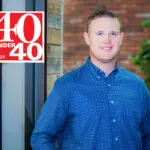 A smiling man in a blue plaid shirt stands indoors by a wooden wall. The 2025 “40 Under 40” logo is displayed in the upper left corner. A brick wall and a green plant are visible in the background.