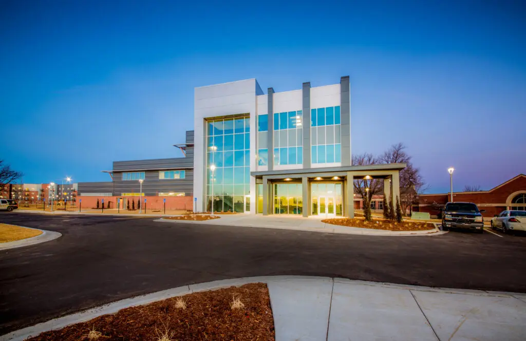 A modern, three-story building with large glass windows is brightly lit at dusk. The surrounding area includes a paved driveway, parked cars, and landscaped grounds with mulch and young trees.