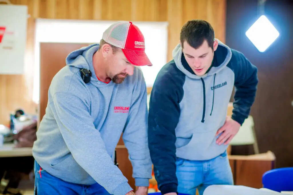 Two men wearing gray hoodies and jeans are standing indoors, looking down and discussing papers or plans on a table. One wears a red cap; both appear focused and engaged in their work.
