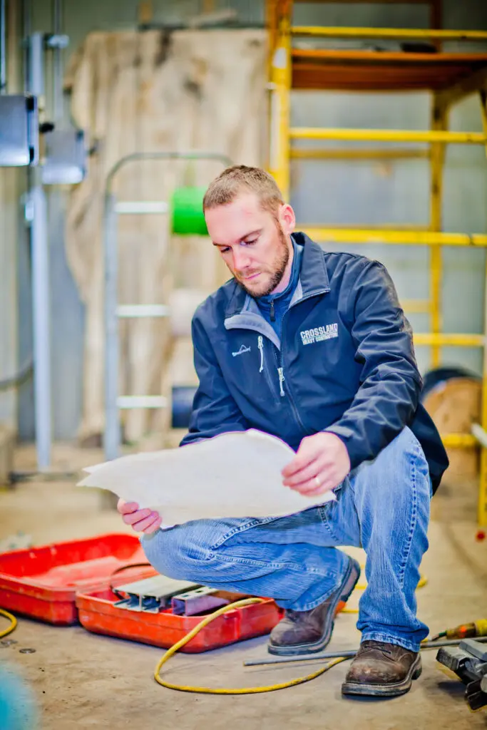 A man in a jacket and jeans kneels indoors on one knee, looking at a large sheet of paper. Tools are visible in an open red toolbox on the floor beside him, with scaffolding and workshop equipment in the background.