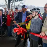 A group of people gather under a pavilion as a man in a suit and two young people wearing bike helmets cut a large red ribbon with oversized scissors, celebrating the opening of a new space or facility.