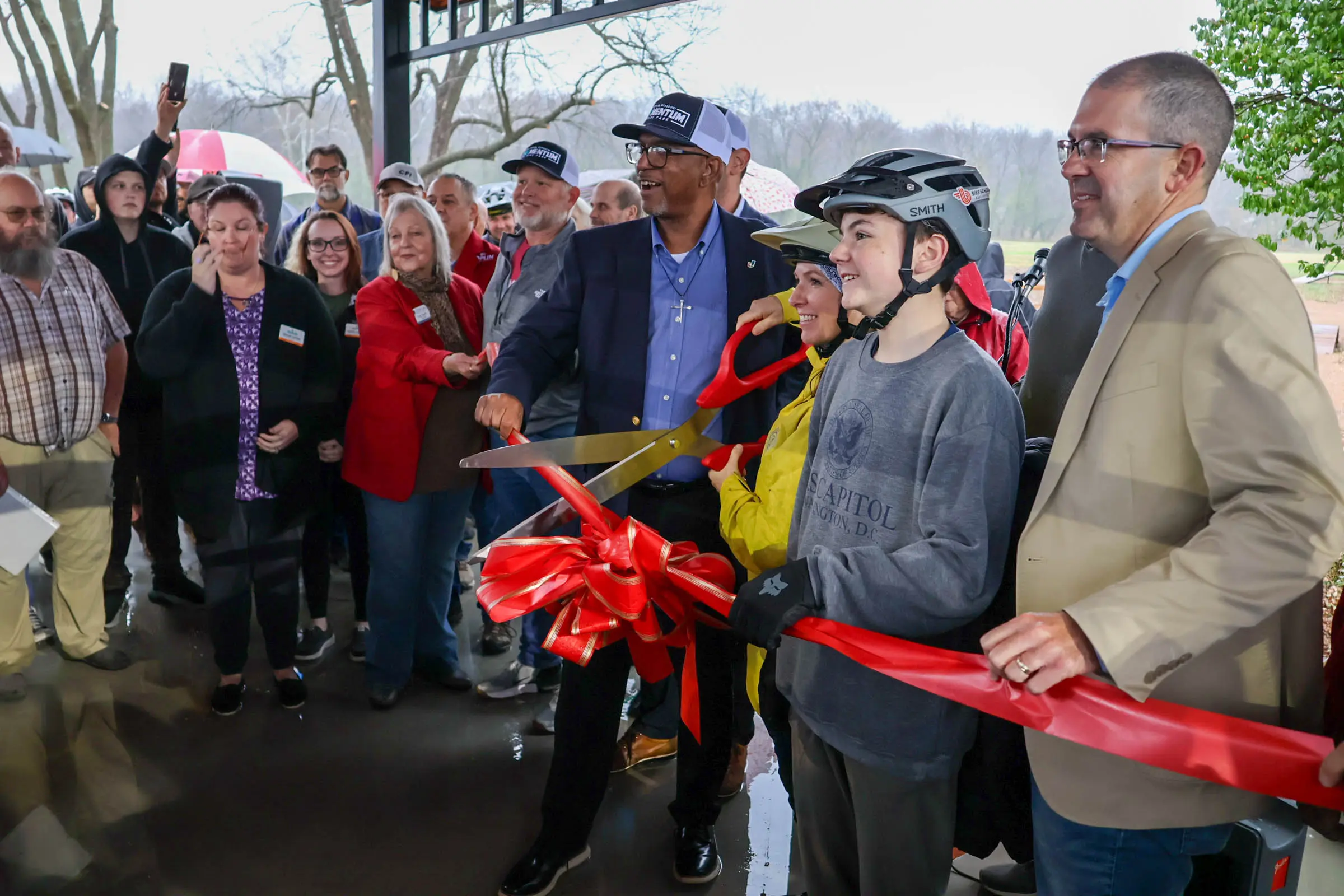 A group of people gather under a pavilion as a man in a suit and two young people wearing bike helmets cut a large red ribbon with oversized scissors, celebrating the opening of a new space or facility.
