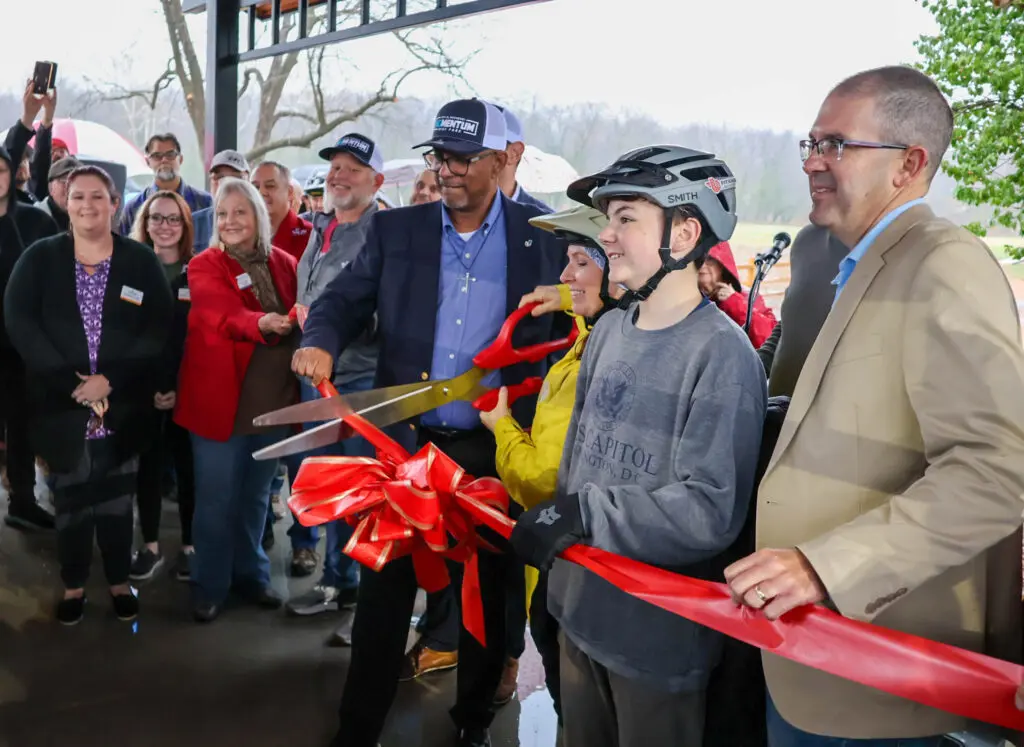 A group of people gathers for a ribbon-cutting ceremony. A smiling boy in a helmet holds a large pair of scissors with two adults, surrounded by onlookers under a covered outdoor area. A red ribbon and bow are in front.