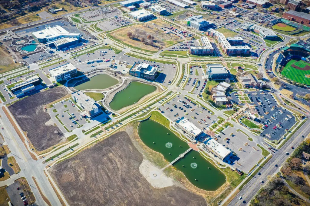 Aerial view of a suburban area with several buildings, parking lots, green spaces, two ponds with fountains, and a baseball field. Roads and sidewalks connect the campus-like complex.