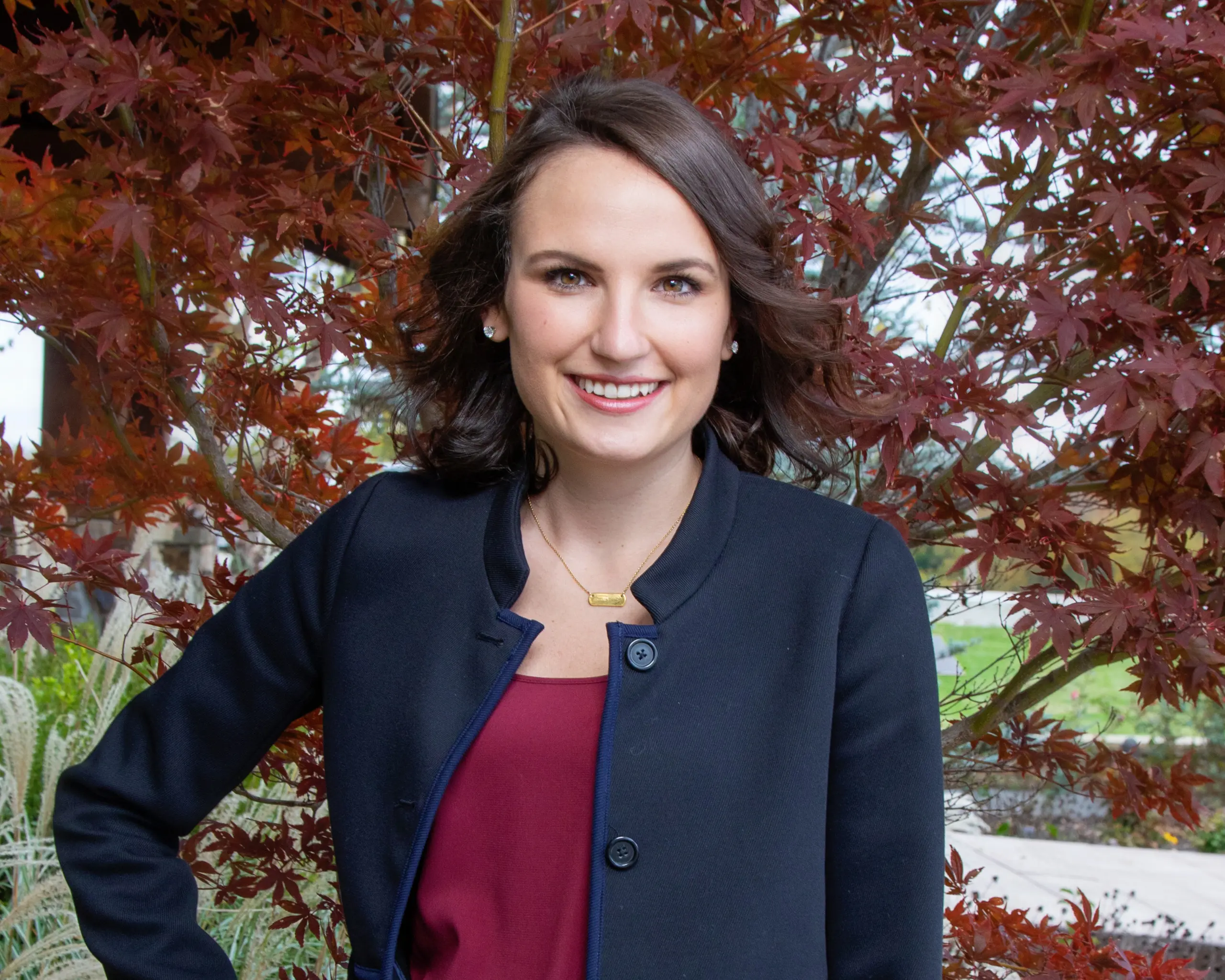 A woman with shoulder-length brown hair, wearing a black blazer over a burgundy top, stands smiling in front of a tree with reddish leaves.