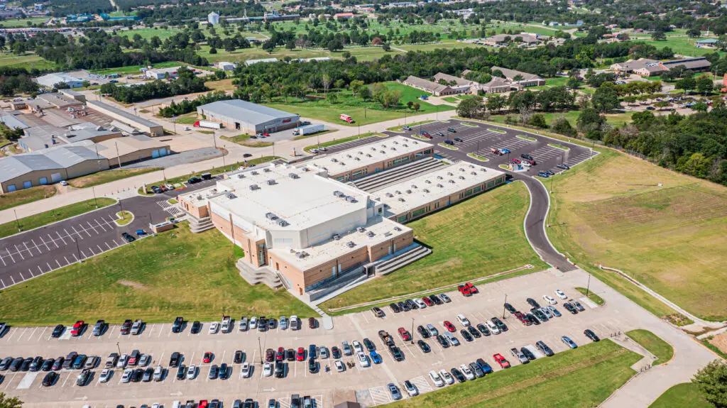 Aerial view of a large modern building with a white roof, surrounded by parking lots filled with cars and green grassy areas, with additional buildings and trees in the background.