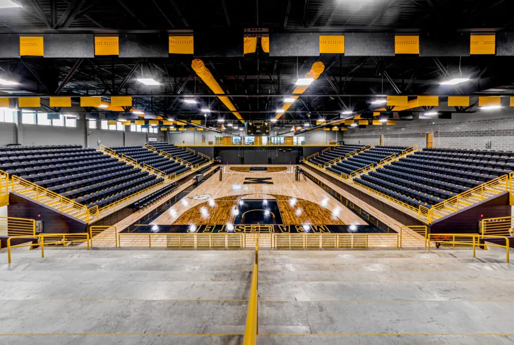 Empty indoor basketball arena with rows of black seats, yellow railings, wooden court with "WEST GEORGIA" written on it, and yellow-and-black banners hanging from the ceiling.
