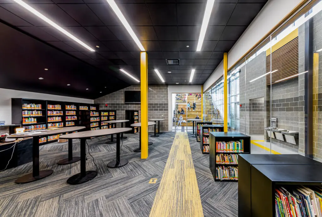Modern library interior with bookshelves, high tables, yellow columns, and a glass wall showing a staircase and seating area. Large windows let in natural light, and water fountains are visible near the entrance.