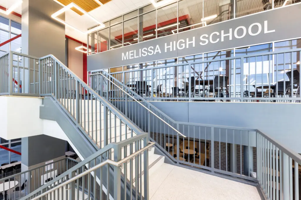 Wide staircase and metal railings inside a modern school building with large windows and a sign reading "MELISSA HIGH SCHOOL" above a glass-walled room on the upper floor.