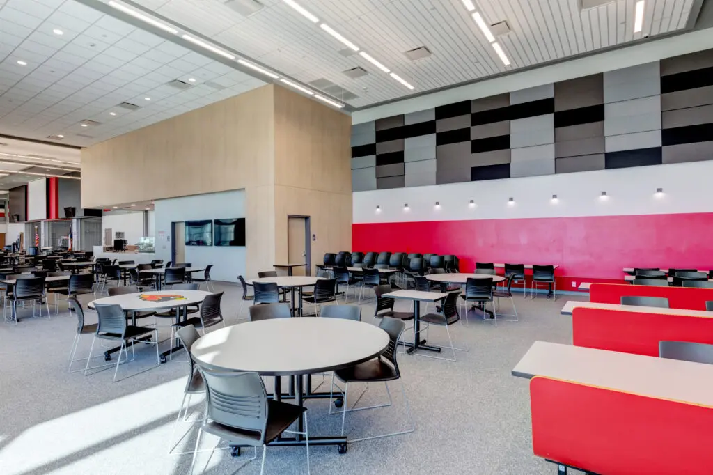 A modern, spacious cafeteria with round tables, black chairs, red and gray booths, and a bold red accent wall beneath geometric gray panels. Bright lighting and large windows create an open, welcoming atmosphere.
