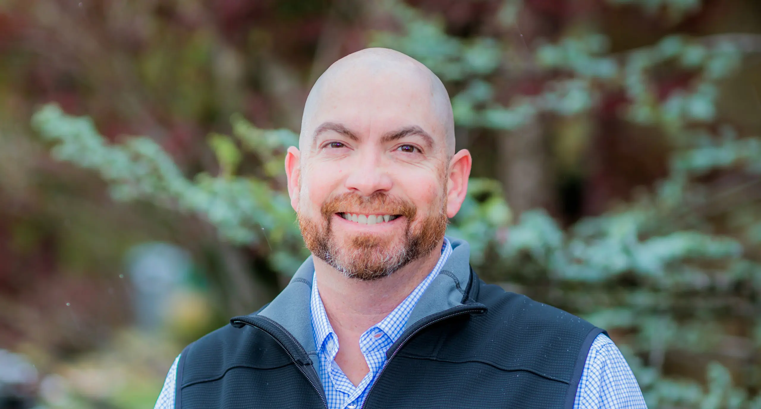 A smiling bald man with a trimmed beard, wearing a blue checkered shirt and a black vest, stands outdoors with blurred green foliage in the background.