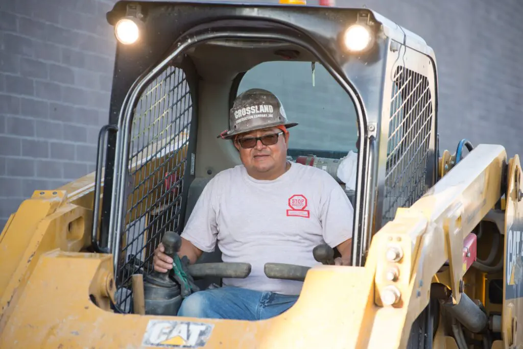A construction worker wearing safety glasses and a hard hat sits inside the cab of a yellow CAT skid steer loader, operating the controls.