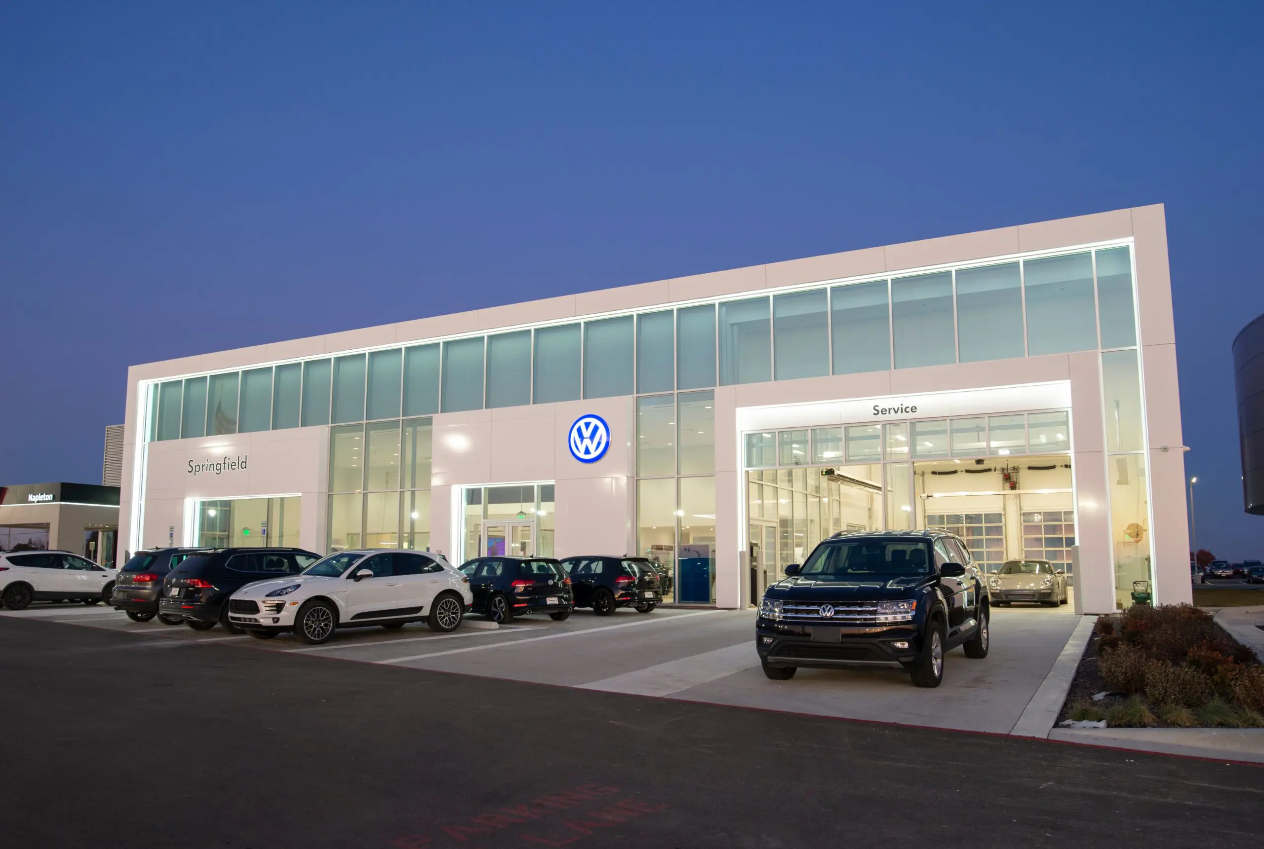 A modern Volkswagen car dealership at dusk, with large glass windows, several parked cars in front, and illuminated signage reading "Springfield" and "Service." The sky is clear and blue.
