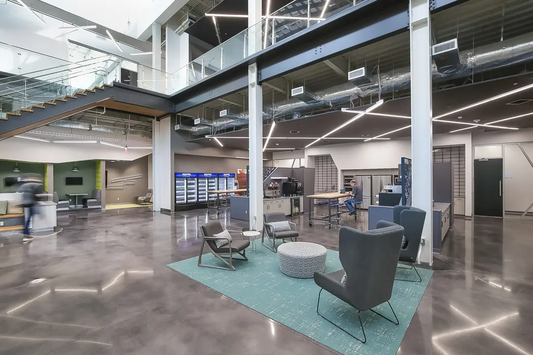 A modern, open workspace with polished concrete floors, lounge chairs on a blue rug, and workstations. There is a staircase, glass railings, and exposed ductwork, creating an industrial yet inviting atmosphere.