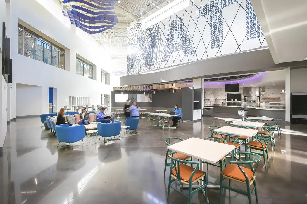 A modern, spacious cafeteria with blue lounge chairs, tables, and people conversing. The floor is polished concrete, and "SIAT" is displayed in large letters on an upper wall. The area is bright with natural light.
