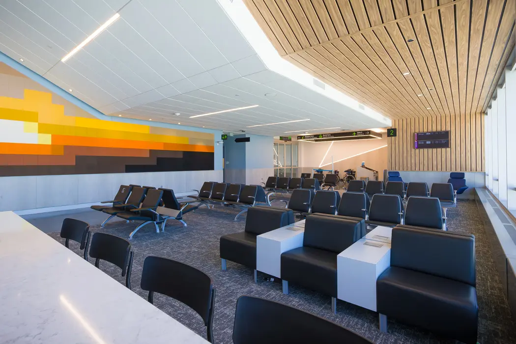 A modern airport waiting area with rows of black seats, white tables, and large windows. A mural with yellow and orange geometric shapes decorates one wall. Digital signs hang above the boarding gates.