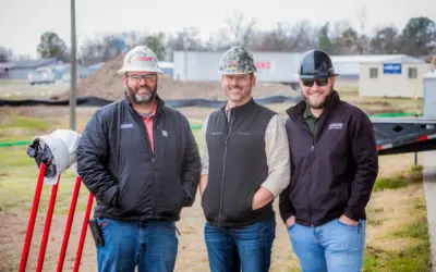 Three smiling men wearing hard hats and jackets stand outdoors at a construction site. There is equipment and dirt in the background, with buildings and trees in the distance.