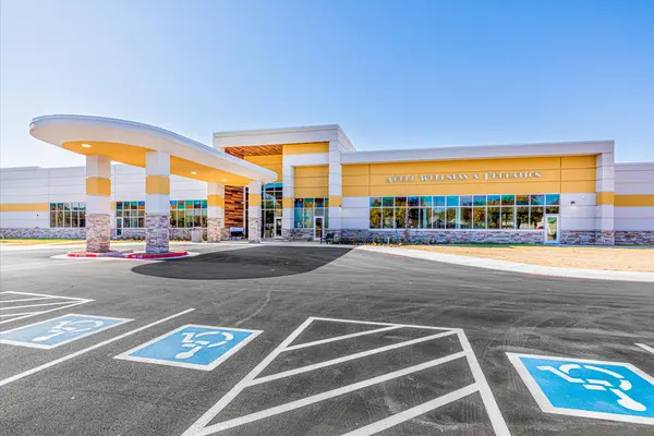 Modern building with large windows and yellow accents, featuring a covered entryway. Several accessible parking spaces with blue wheelchair symbols are in front of the entrance. Clear sky is visible above.